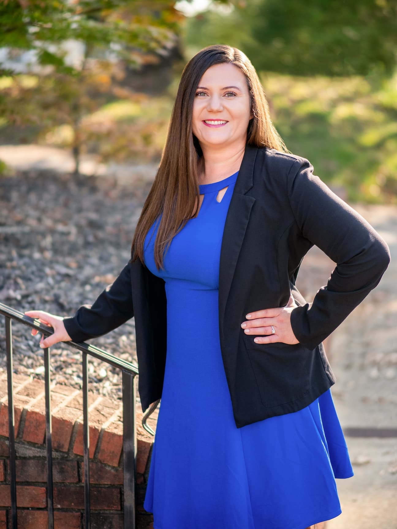 An attorney, Mirsada Kanwisher, wearing a blue dress, facing the camera, and smiling while standing outdoors holding a railing.